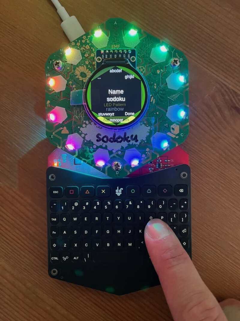 The small Hexpansion keyboard by davedarko and sodoku. It is the same size as the whole Tildagon badge, and links to the bottom two edges of the hexagon. Someone's finger is typing their name which appears on the screen.