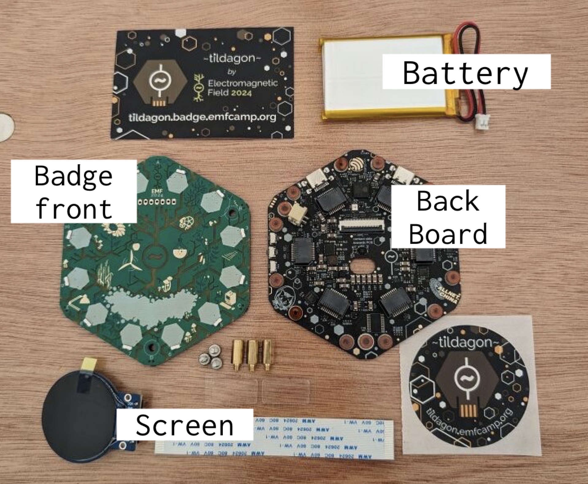 These are the parts of the badge (last time's design for 2024 shown). There is a silver flat battery with a JST connector, a hexagonal plate labelled 'badge front', a black hexagonal plate with microcontroller chips and visible components labelled 'back board', a small round screen with a screen protector on, and also a flat ribbon cable, some standoffs and screws, and a sticker and business card that say Tildagon. It was the contents of a full badge pack in 2024. The design for 2026 has not been released yet.